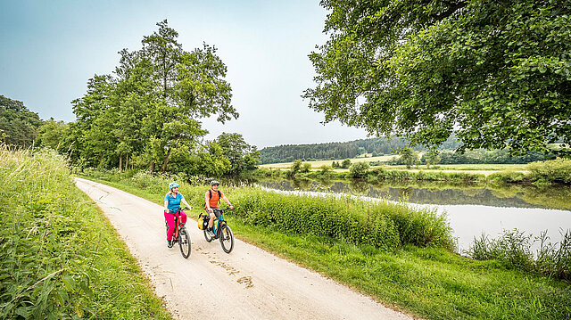 Zwei Radfahrer radeln an einem Fluss entlang.