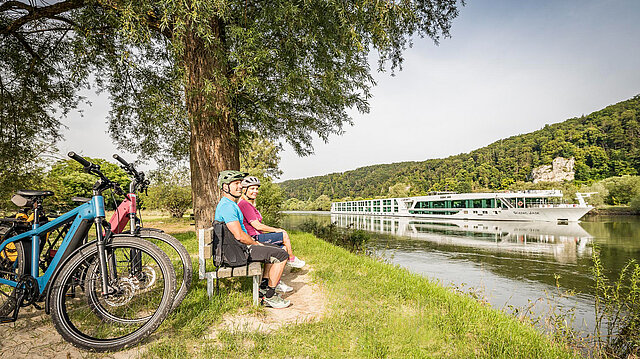 Zwei radfahrer sitzen auf einer Bank und schauen auf ein vorbeifahrendes Flussschiff